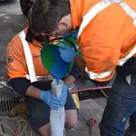 two workers helping each other in pouring blue coloured chemical into a Pipe